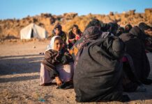 A young girl in traditional clothing sitting among a group of women in a refugee camp