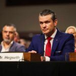 Maximum Violence Teased — What’s The Endgame? A man in a blue suit and red tie sitting at a hearing table with a nameplate