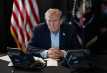A serious-looking man seated at a table with phones and an American flag in the background