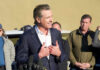 Politician speaking at a press conference with a group of people in the background