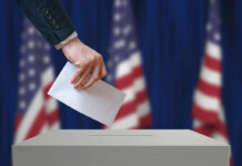 Trump Overhauls Federal Election Agency A hand placing a ballot into a ballot box with American flags in the background