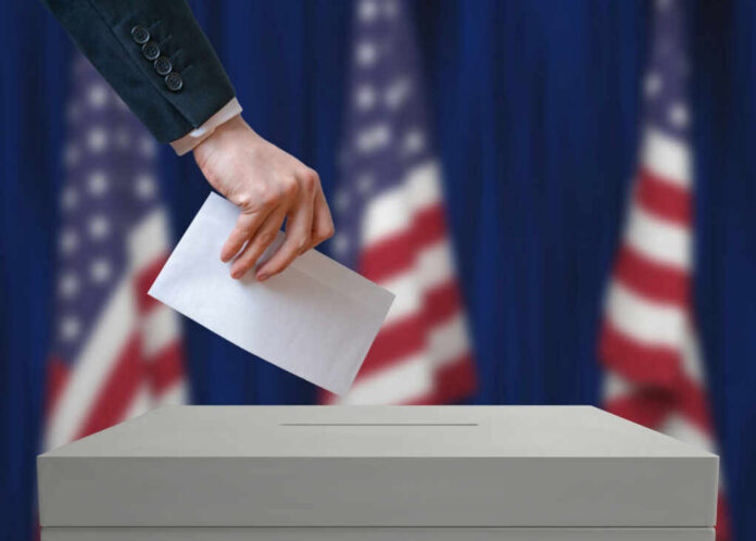 shutterstock426222397jpg A hand placing a ballot into a ballot box with American flags in the background
