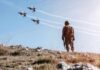 Soldier standing on rocky hillside watching fighter jets fly overhead