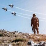 U.S.-Saudi Strike Escalates Regional Conflict Soldier standing on rocky hillside watching fighter jets fly overhead