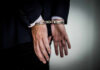 Fugitive Doctor Captured After Two Decades A person in a suit with hands cuffed behind their back