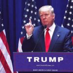 man in suit speaking at podium with multiple American flags