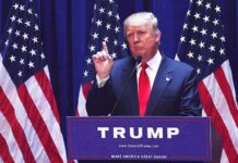 man in suit speaking at podium with multiple American flags