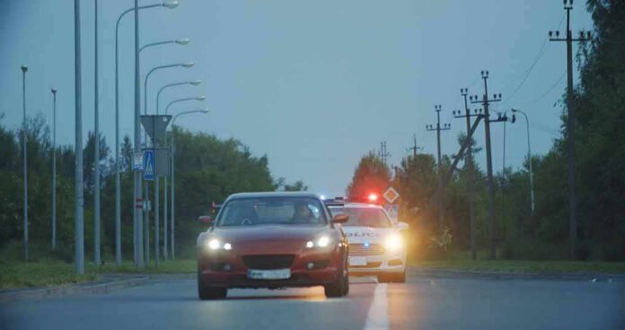 shutterstock_1642255009 red car driving on road with police car behind using flashing lights