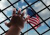 Hand reaching through fence toward blurred American flag