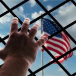 Hand reaching through fence toward blurred American flag