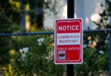 The Flock Camera Debate Just Escalated Red notice sign on a black pole in a leafy outdoor area