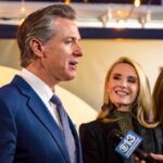 Man in a blue suit speaks to reporters while two women stand beside him