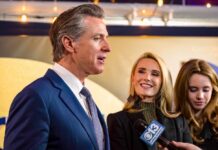 Man in a blue suit speaks to reporters while two women stand beside him