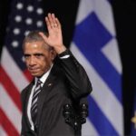 Barack Obama waves on stage with flags in the background